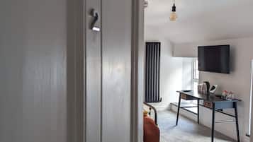 Une pièce avec une porte blanche, un radiateur noir, une table noire avec un téléviseur, une chaise et une ampoule suspendue.
