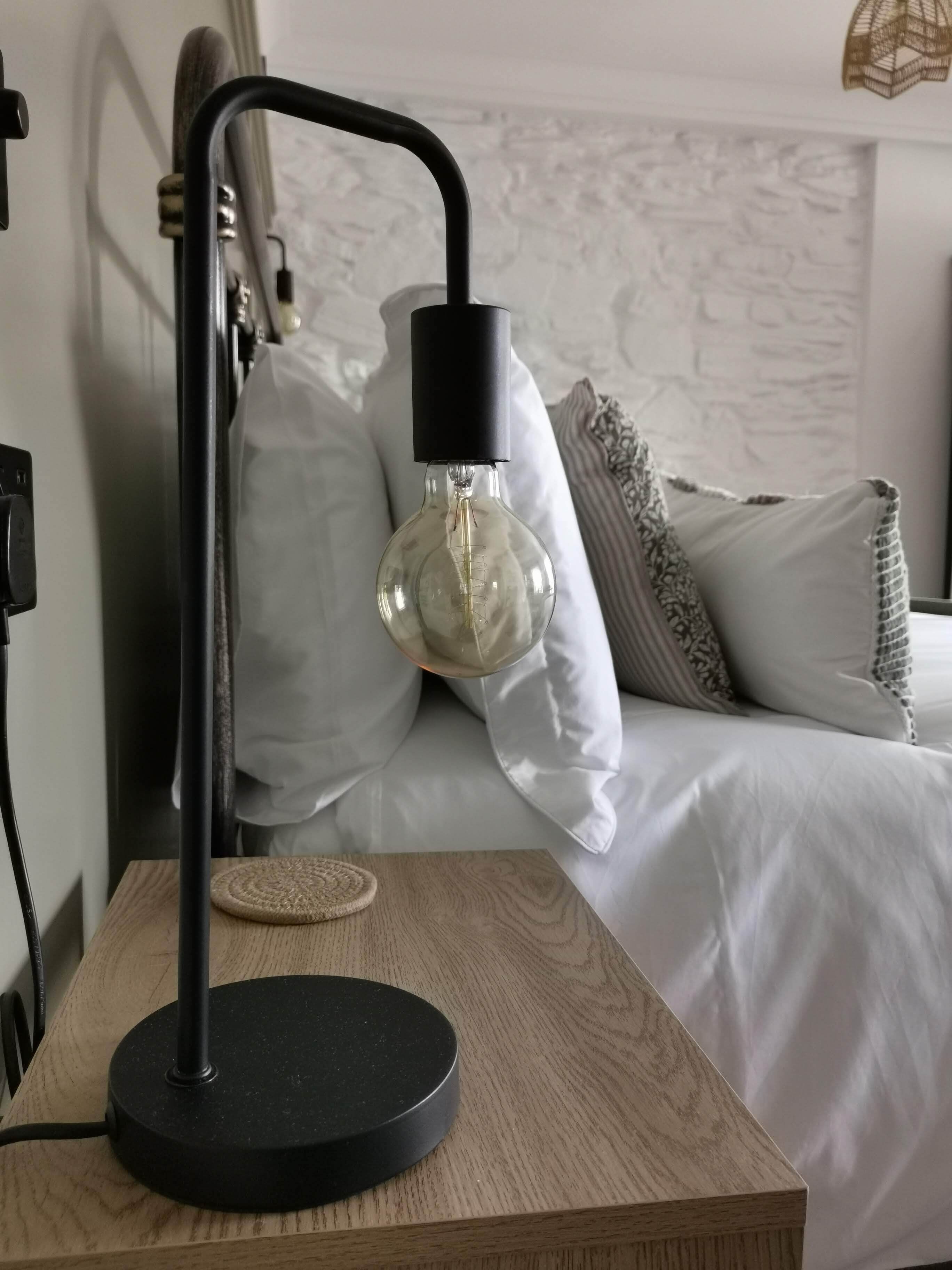 Quarto moderno com mesa de cabeceira de madeira, abajur preto e cama com roupa de cama branca e almofadas estampadas.