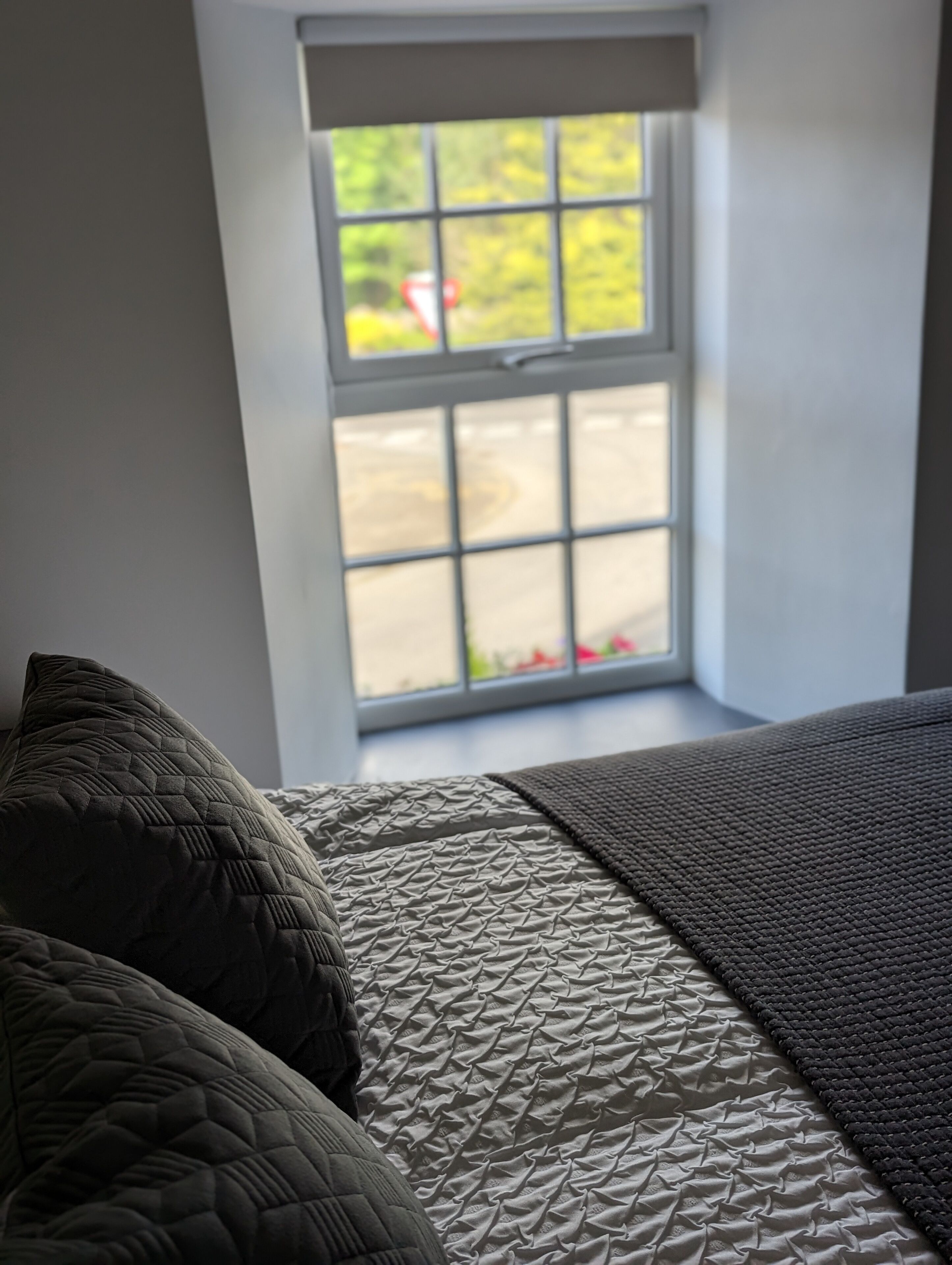 Een slaapkamer met een bed, kussens en een raam met uitzicht op groen en een rood voorwerp buiten.