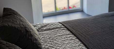 Een slaapkamer met een bed, kussens en een raam met uitzicht op groen en een rood voorwerp buiten.