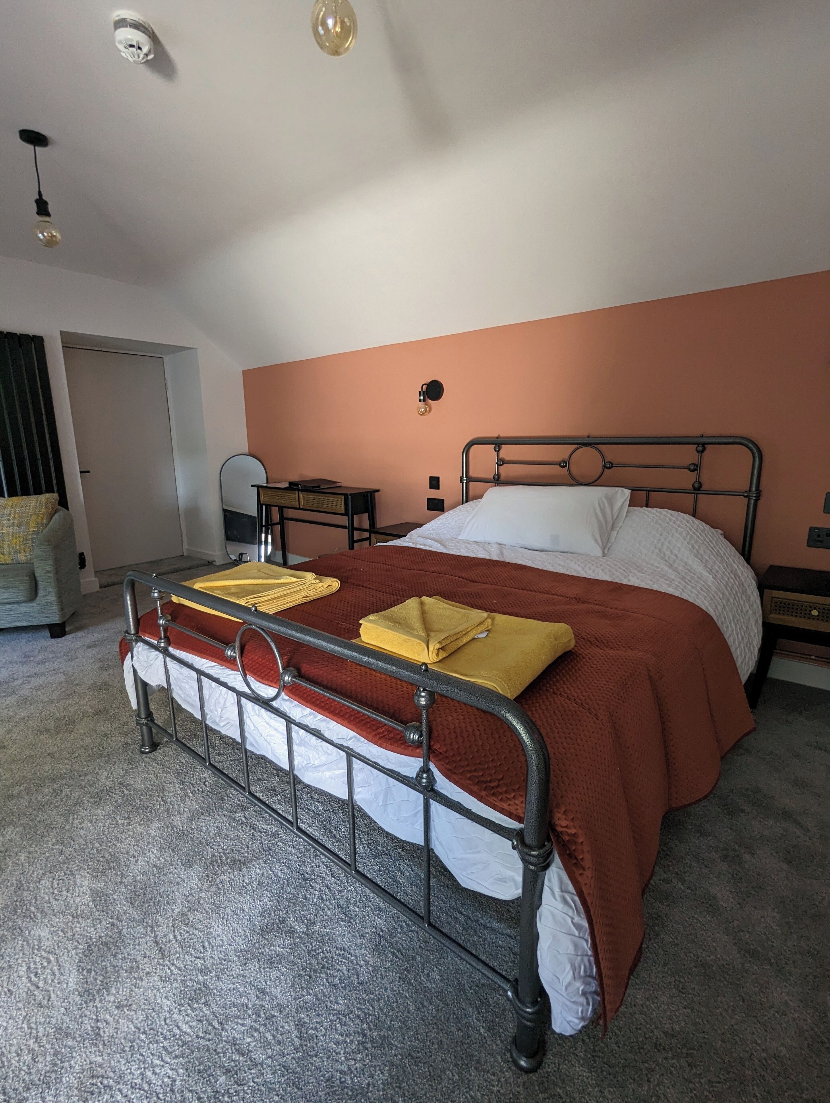 Ein Schlafzimmer mit einem Metallbettgestell, einer roten und weißen Bettdecke, einem gelben Handtuch und einem kleinen Nachttisch mit einer Lampe.