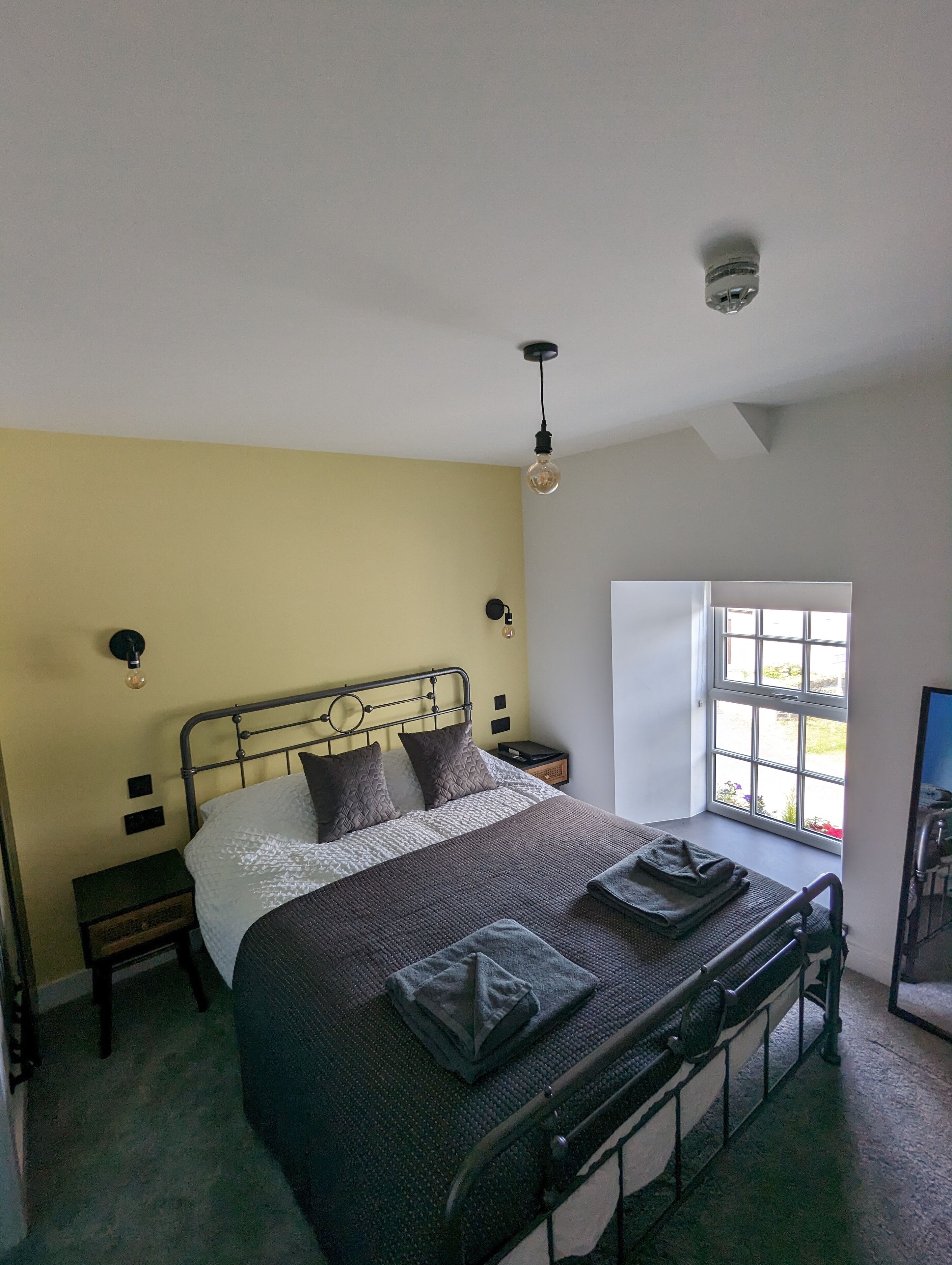 A bed with a metal frame, a bedside table, a lamp, and a window with a view of the outdoors.