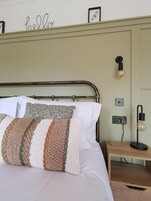 Una cama bien hecha con cabecera metálica, una lámpara de pared y una mesita de noche con lámpara.