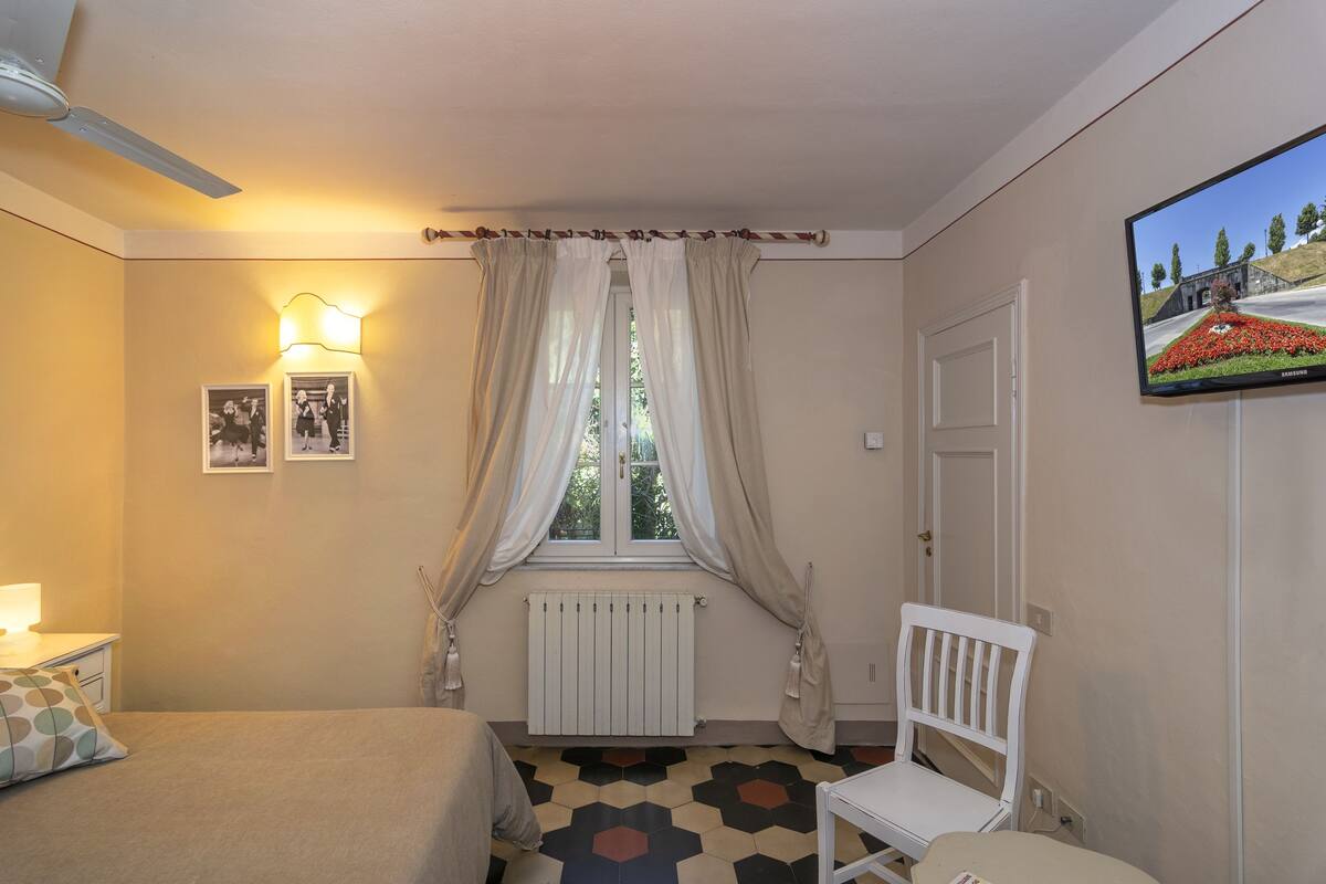 Une chambre à coucher comprenant un lit, une chaise, un radiateur, une fenêtre avec des rideaux, un téléviseur fixé au mur et un luminaire fixé au mur.