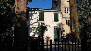 A courtyard with a white building featuring green shutters, surrounded by a black fence, and potted plants. - Corte Meraviglia - Relais (Lucca)