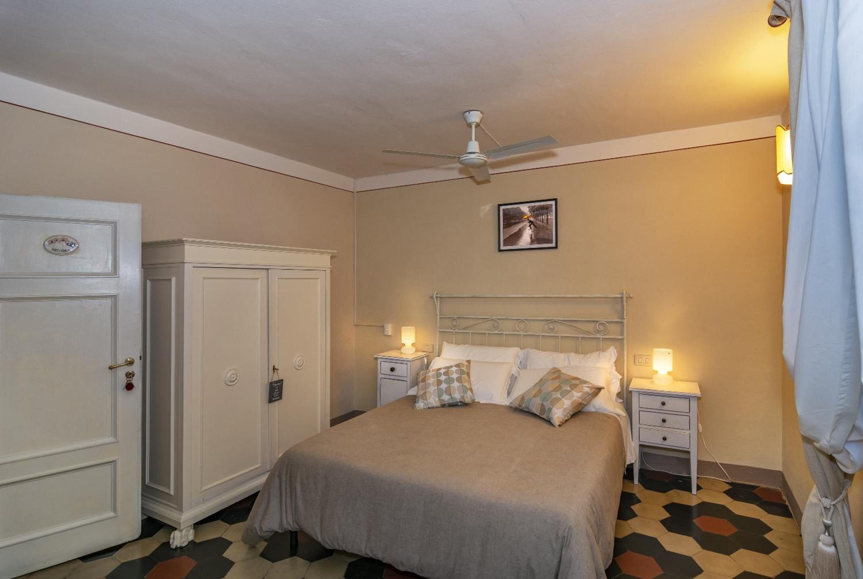 A bedroom with a bed, two bedside tables with lamps, a ceiling fan, a wardrobe, and a door.