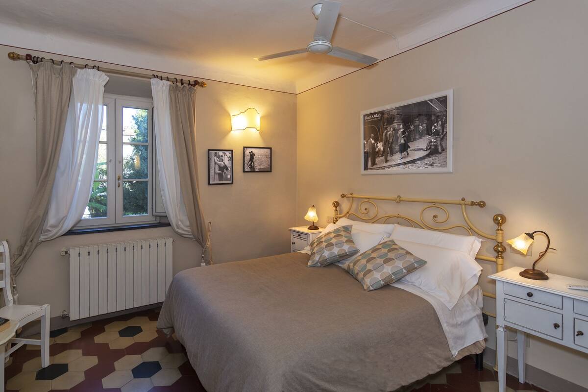 Une chambre à coucher comprenant un lit, une table de chevet, une chaise, un radiateur, une fenêtre avec des rideaux, un ventilateur de plafond et des lampes fixées au mur.