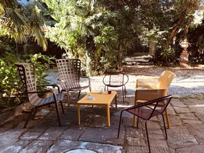 An outdoor seating area with wicker chairs and a table, surrounded by lush greenery and a stone pathway. - Corte Meraviglia - Relais (Lucca)