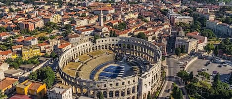 An aerial view of a historic city with a prominent circular building featuring an open central area, surrounded by dense urban development and greenery.