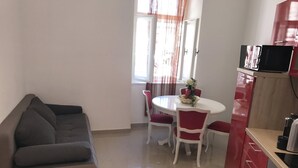 A modern living room with a grey sofa, a round dining table with red chairs, and a kitchen area with red cabinets and a black appliance. - Pula Antic Rooms in Center- Adults only (Pula)