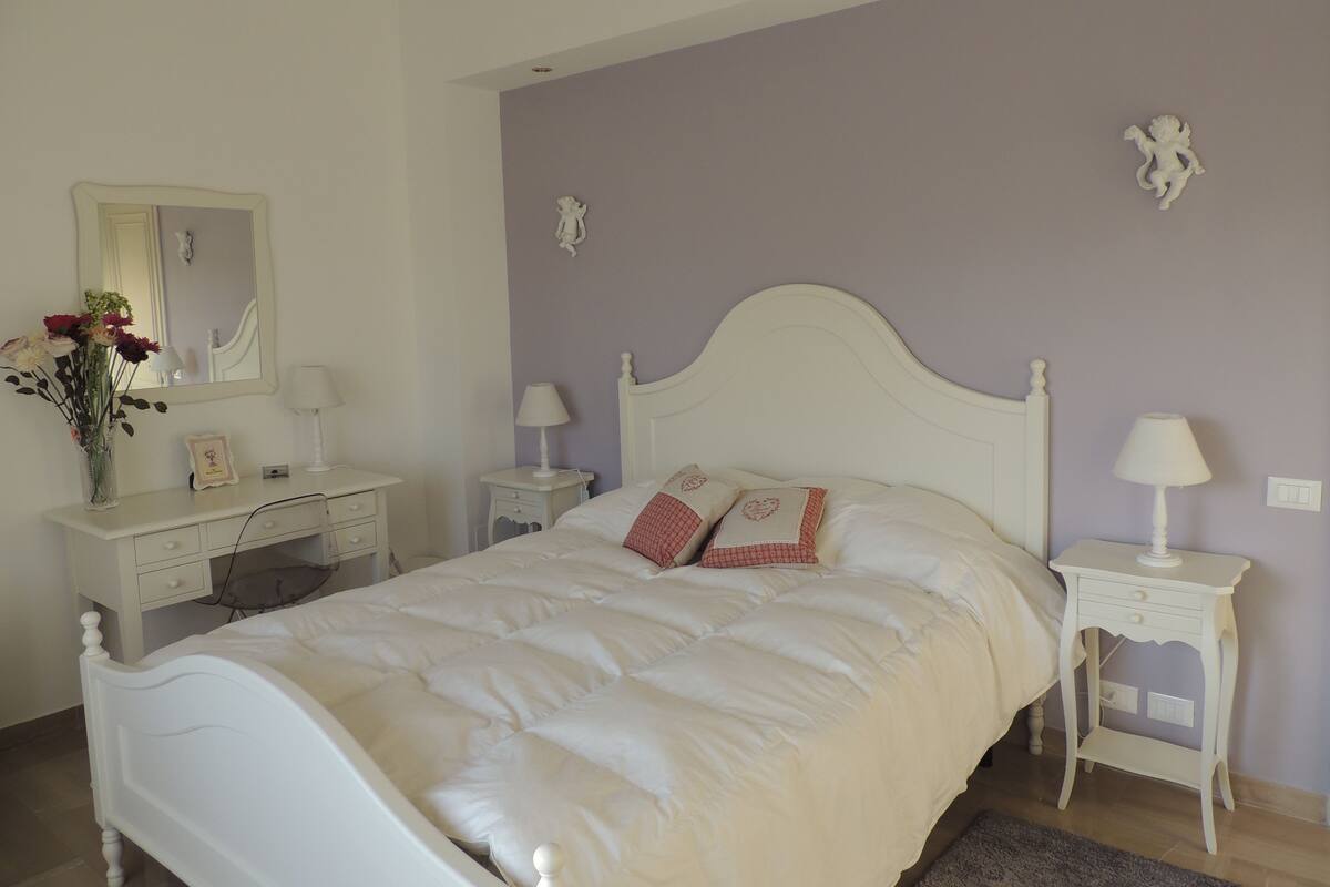 A bedroom with a white bed, a dresser, a nightstand, a mirror, and a wall-mounted decorative piece.