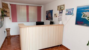 Un mostrador de recepción con detalles de madera, un amplio ventanal con cortinas rosadas y diversos carteles en las paredes.
