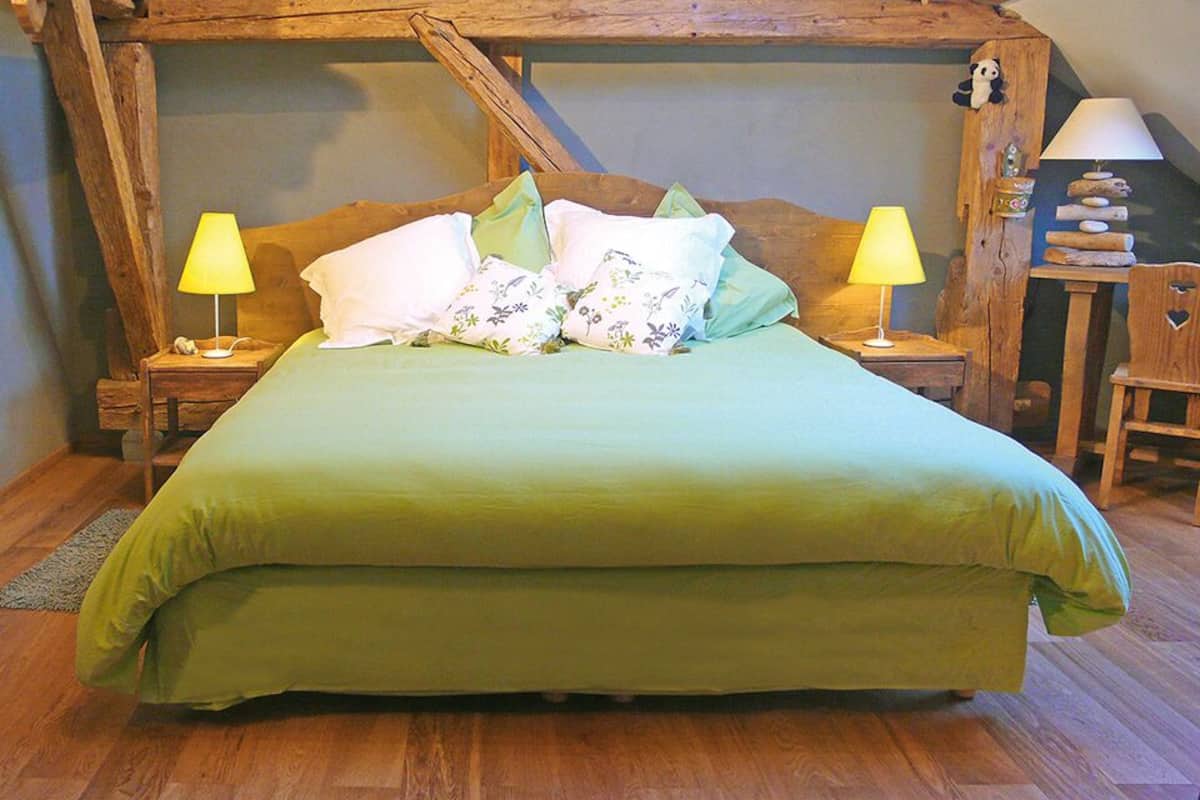 A bed with a green bedspread and floral pillows, two bedside lamps, and a wooden headboard in a rustic room.