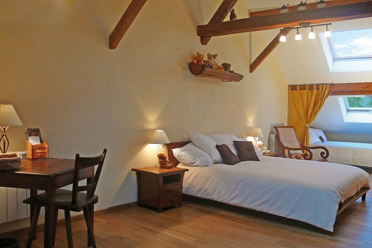 A bedroom with a large bed, a smaller bed, a desk with a chair, a nightstand with a lamp, and a window with curtains.