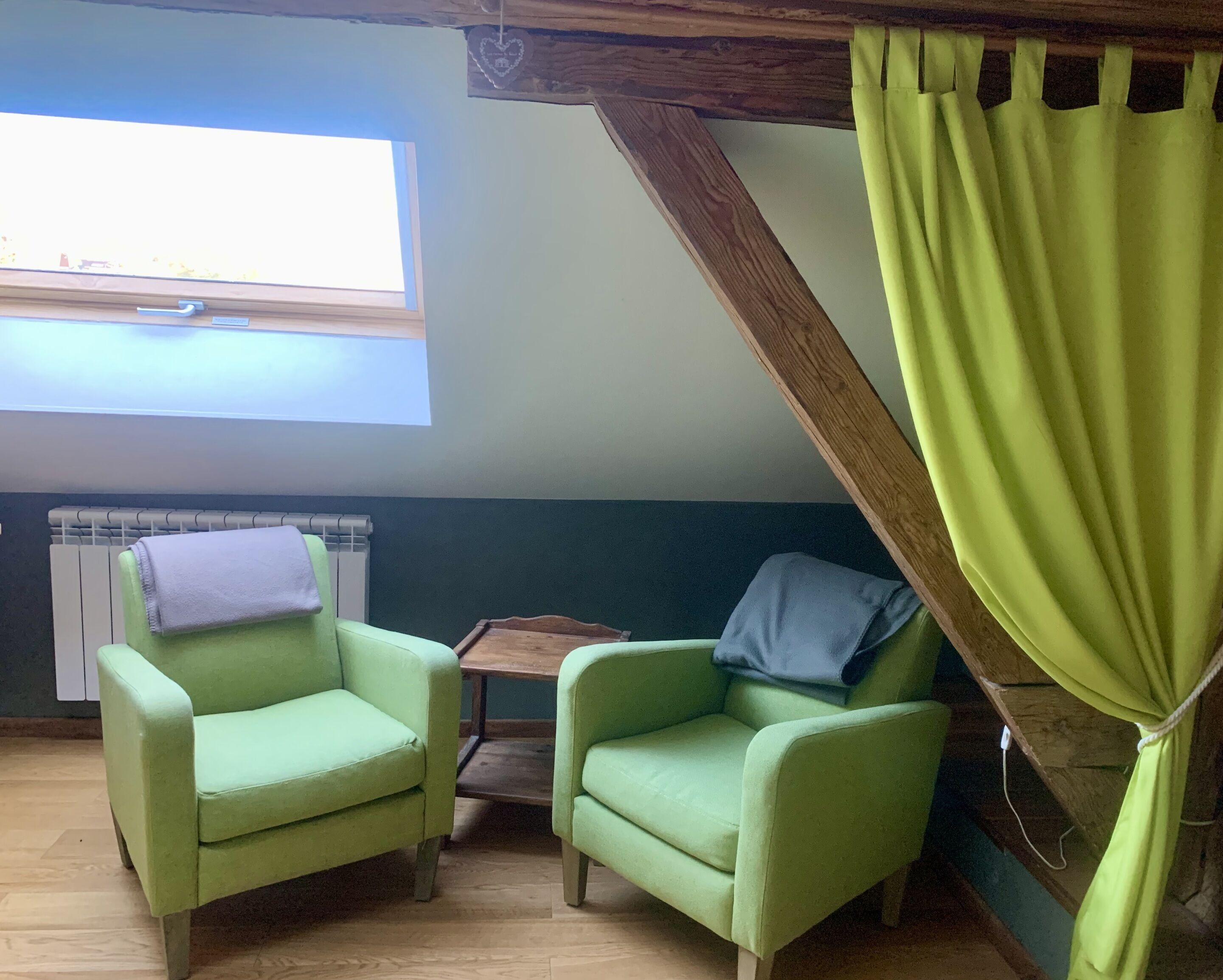 A cozy room with two green armchairs, a small wooden table, a radiator, and a window with a view.