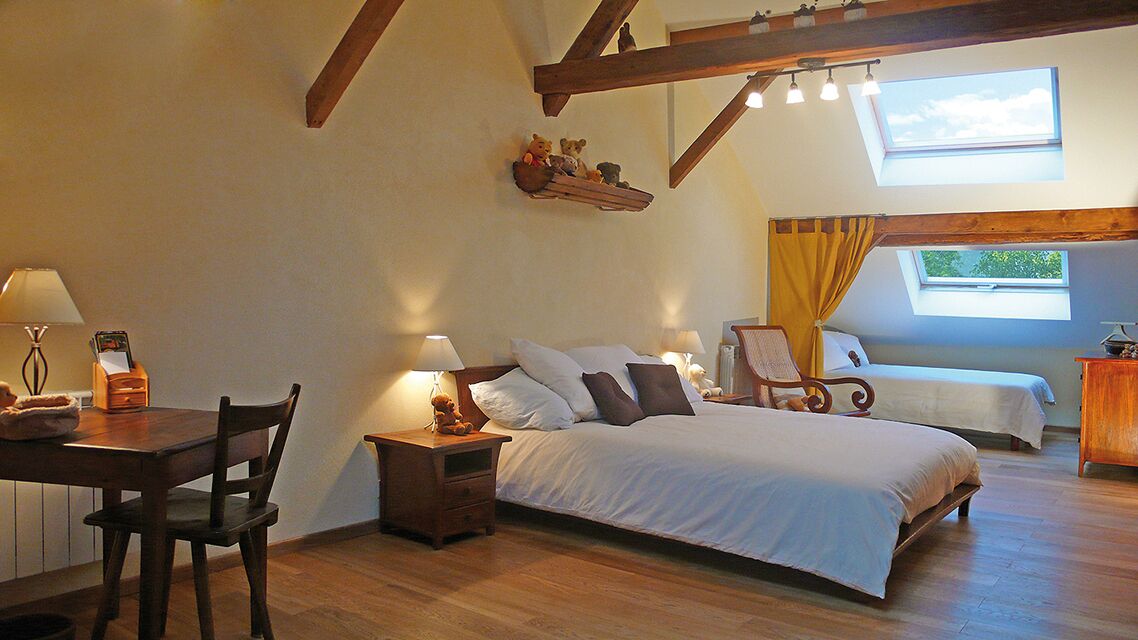 Une chambre confortable avec un grand lit, un plus petit lit, un bureau avec une chaise et une table de chevet avec une lampe. Il y a une fenêtre donnant sur les arbres et un puits de lumière au-dessus du lit.