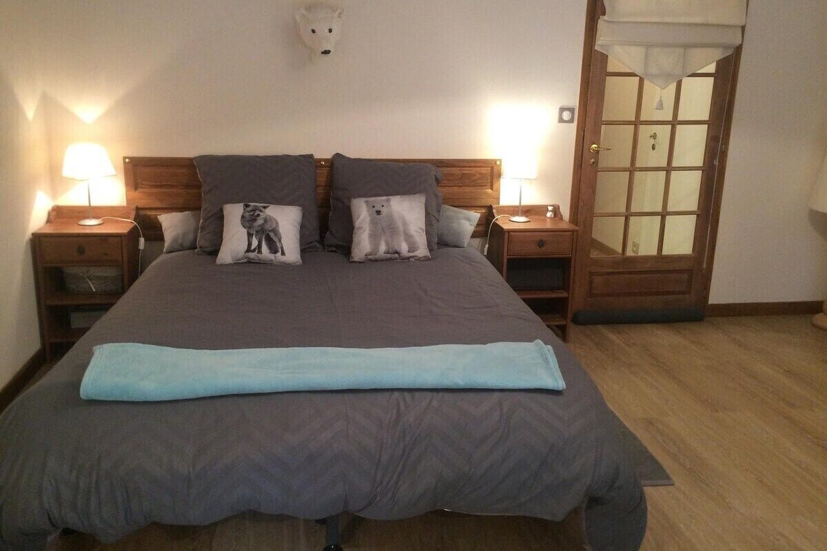 A bedroom with a large bed, two bedside tables, a wooden headboard, a wall-mounted light, a door with glass panels, and a small shelf.