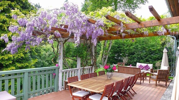 Een houten terras met een pergola bedekt met paarse bloemen, een eettafel met stoelen, en een zithoek met een bank en een parasol.