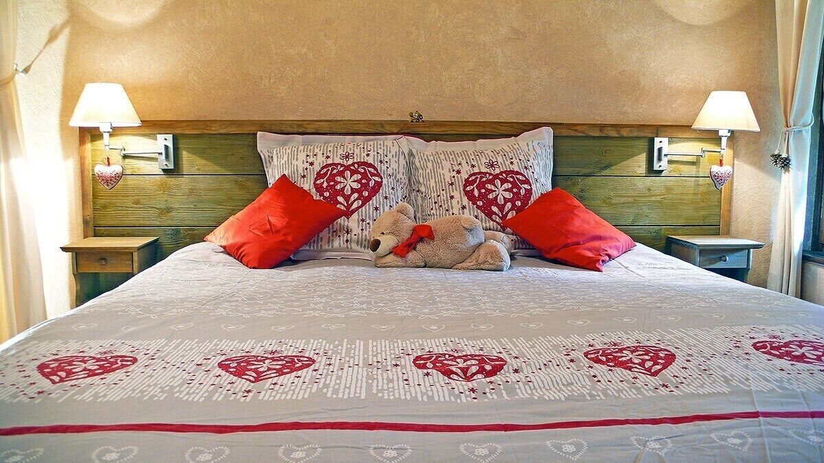 A bed with a heart-patterned bedspread, two red pillows, and a teddy bear.