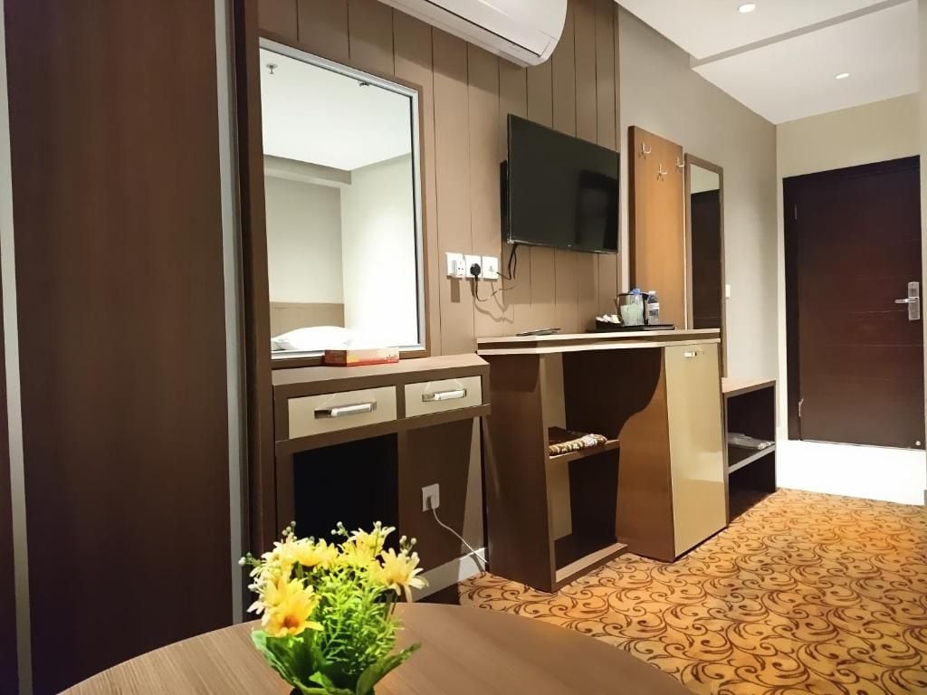 Habitación de hotel con un escritorio de madera, un televisor de pantalla plana montado en la pared, un espejo y una pequeña mesa con un jarrón de flores.