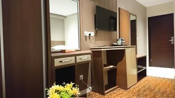 Habitación de hotel con un escritorio de madera, un televisor de pantalla plana montado en la pared, un espejo y una pequeña mesa con un jarrón de flores.