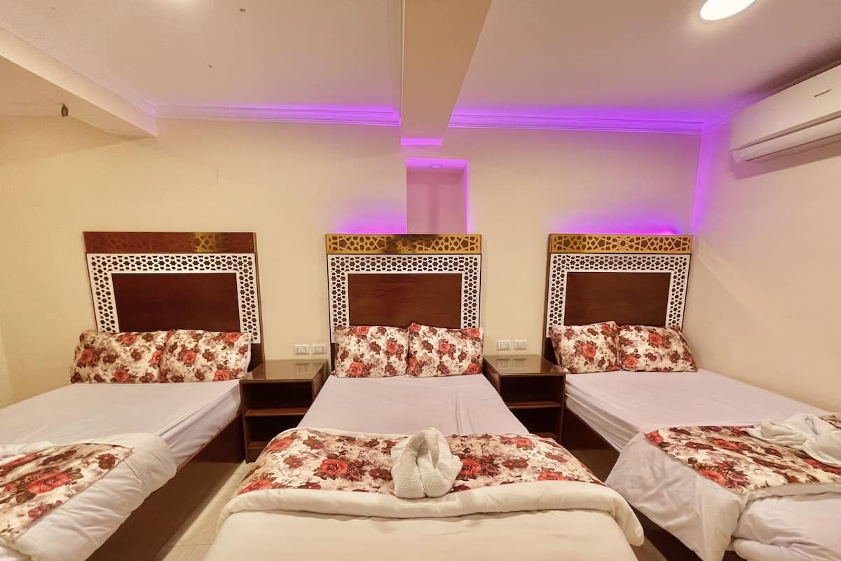 A hotel room with three single beds, each with a floral-patterned headboard and matching pillows. There is a central nightstand with a lamp, and an air conditioning unit mounted on the wall.
