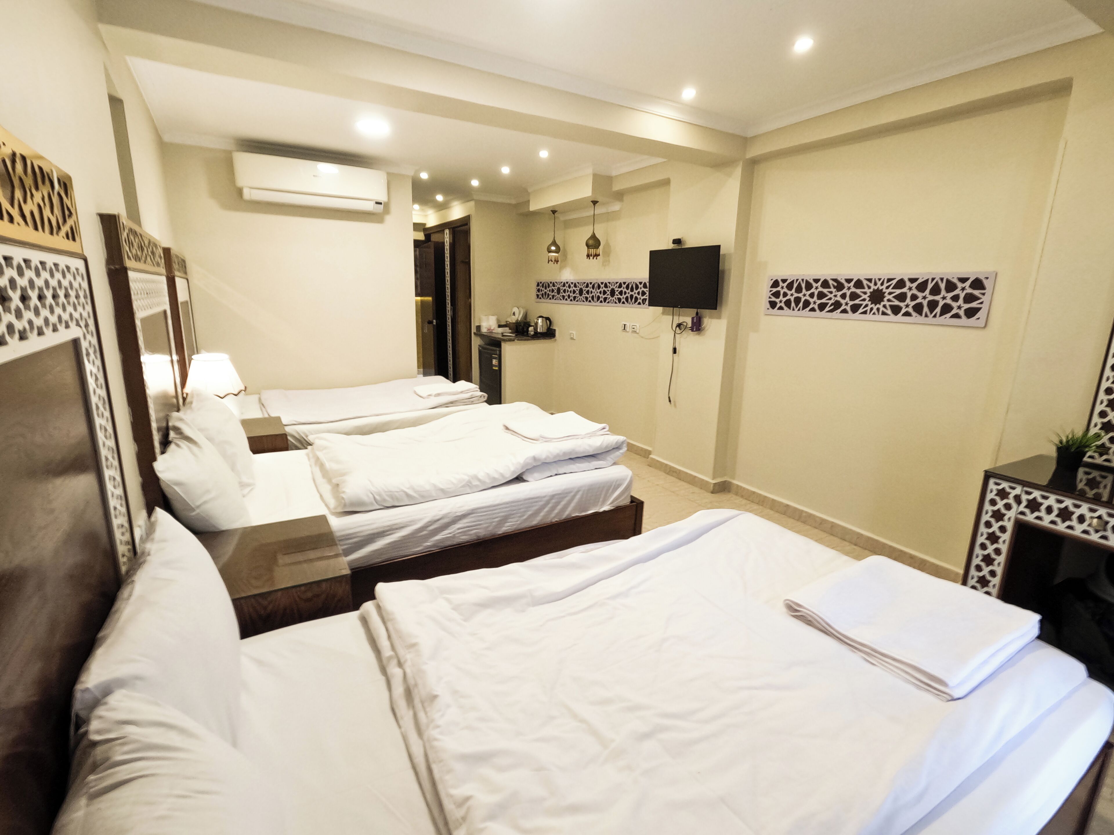 A hotel room with three single beds, a flat-screen TV mounted on the wall, and a small kitchenette area.