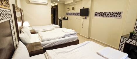 A hotel room with three single beds, a flat-screen TV mounted on the wall, and a small kitchenette area.