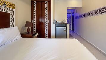 Habitación de hotel con cama, escritorio con lámpara, minibar y puerta que da a otra habitación.