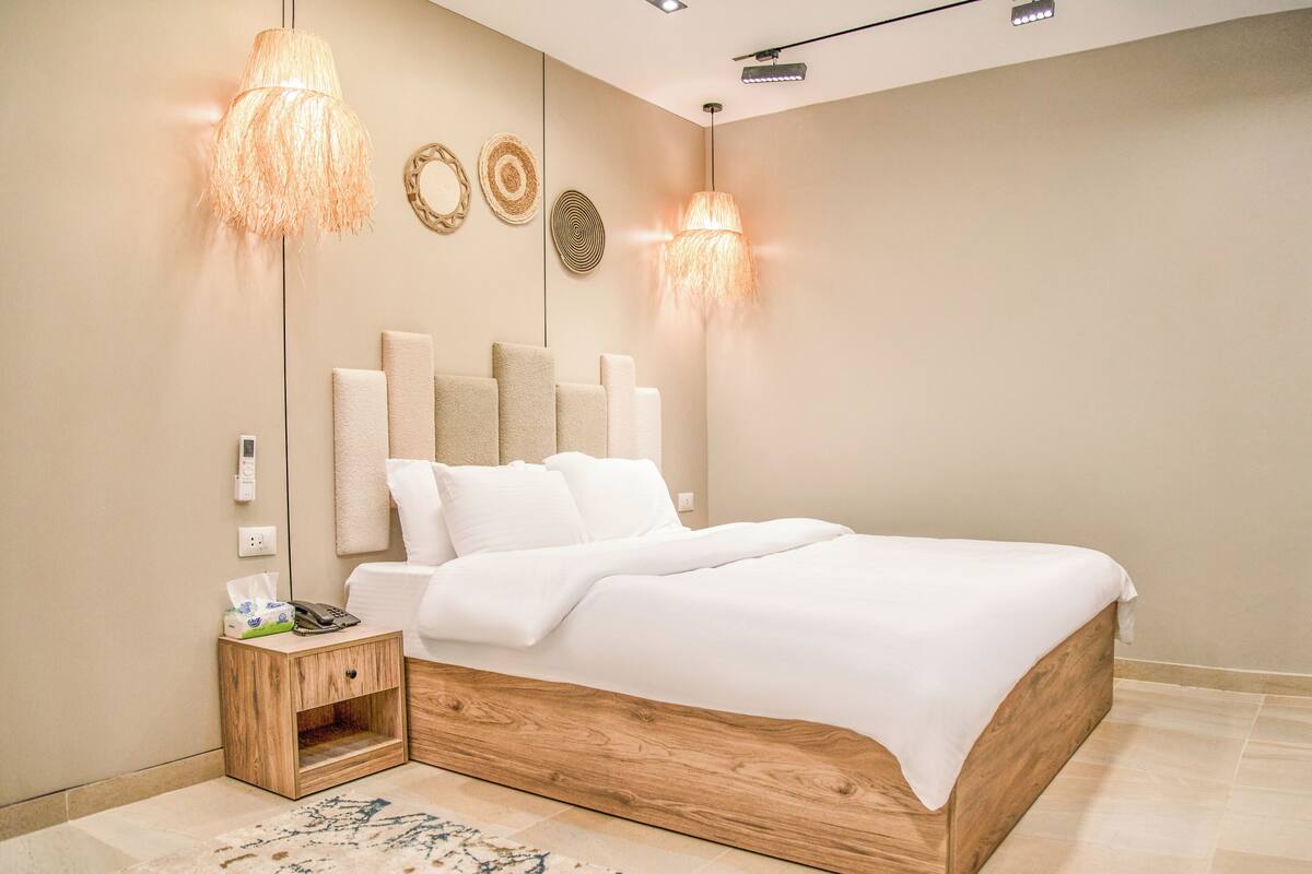 A modern bedroom with a wooden bed, two bedside lamps, a bedside table with a phone and tissues, and a wall-mounted light fixture.