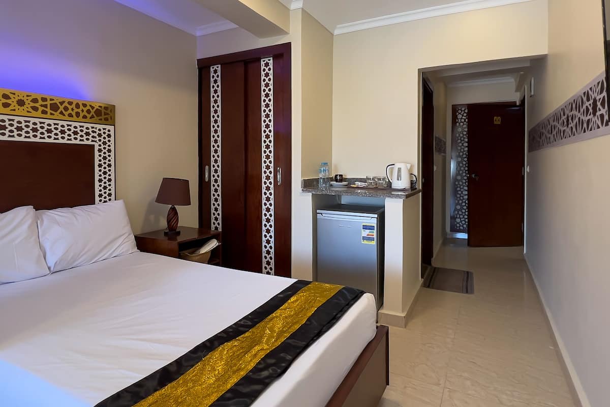 A hotel room with a large bed, a desk with a lamp, a refrigerator, and a minibar.