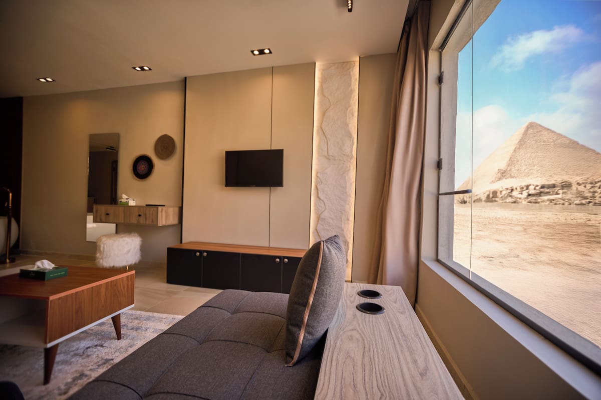 A modern living room with a large window, a flat-screen TV, a wooden coffee table, and a sofa with a cushion.