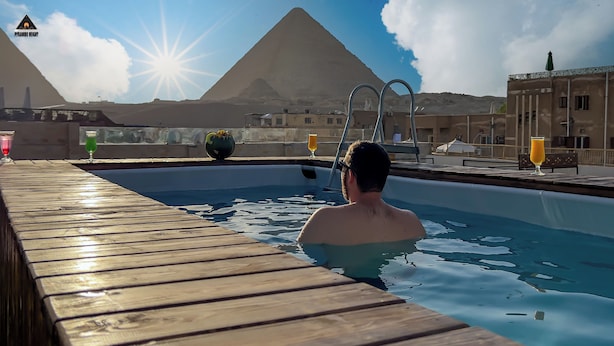 Un área de piscina con cubierta de madera, una persona en el agua y una pirámide al fondo.