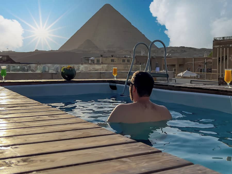 Un área de piscina con cubierta de madera, una persona en el agua y una pirámide al fondo.