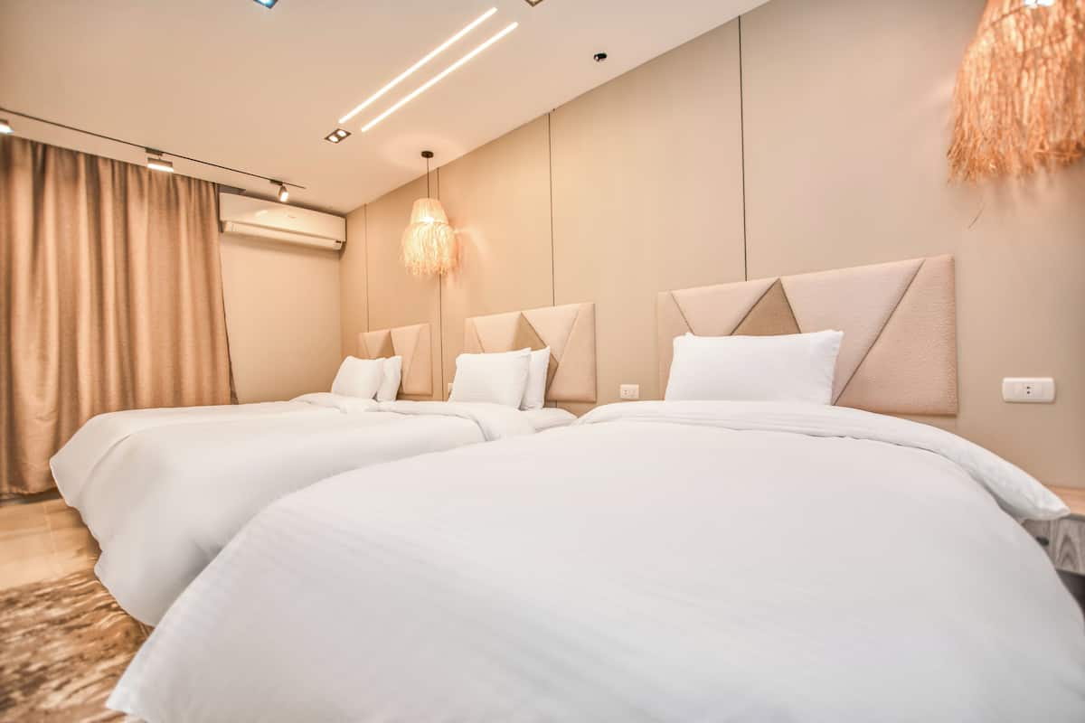 A modern hotel room with two beds, a large window with curtains, a hanging light fixture, and an air conditioning unit.