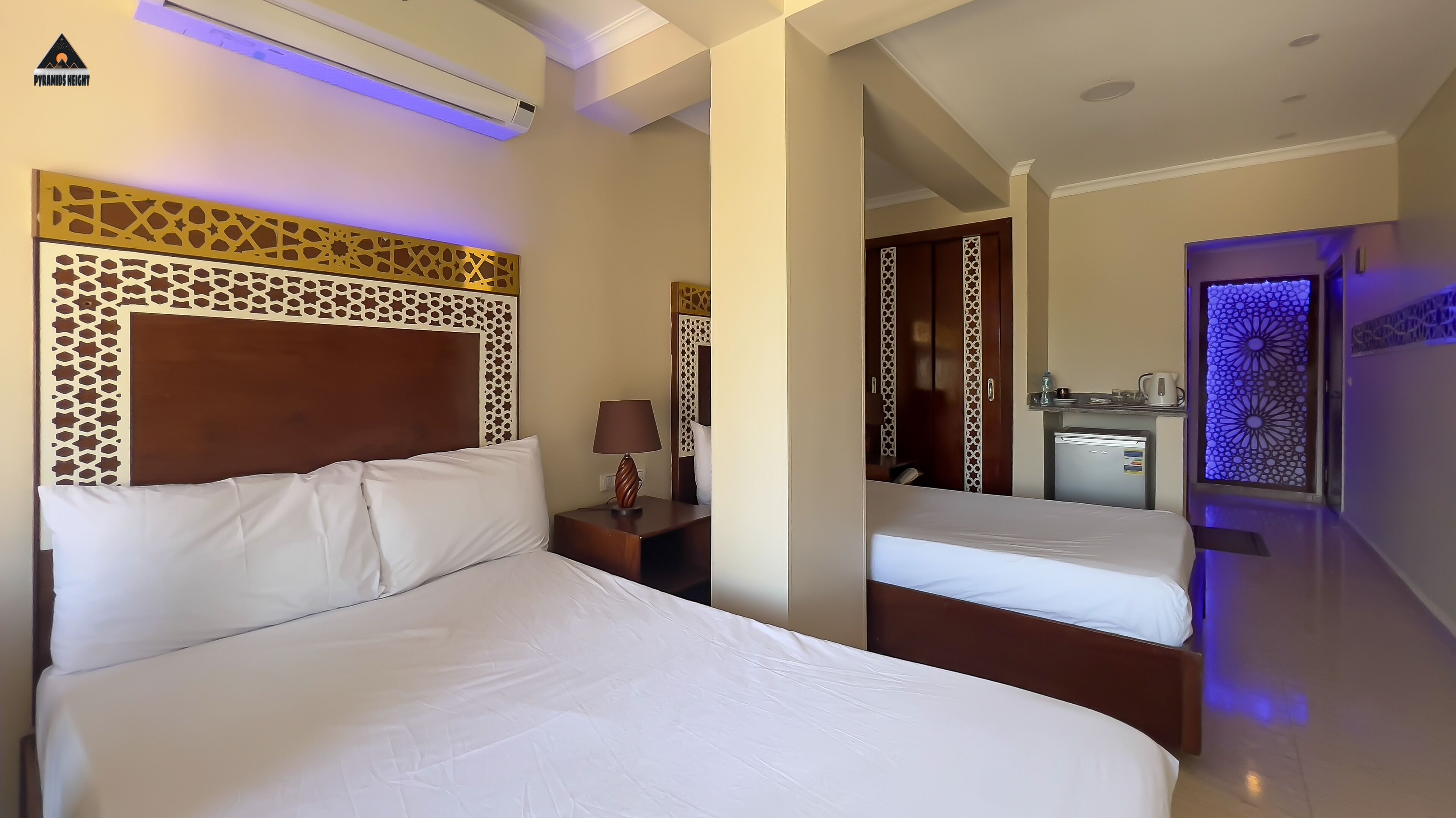 A hotel room with a large bed, a bedside table with a lamp, a wardrobe, and a kitchenette area.