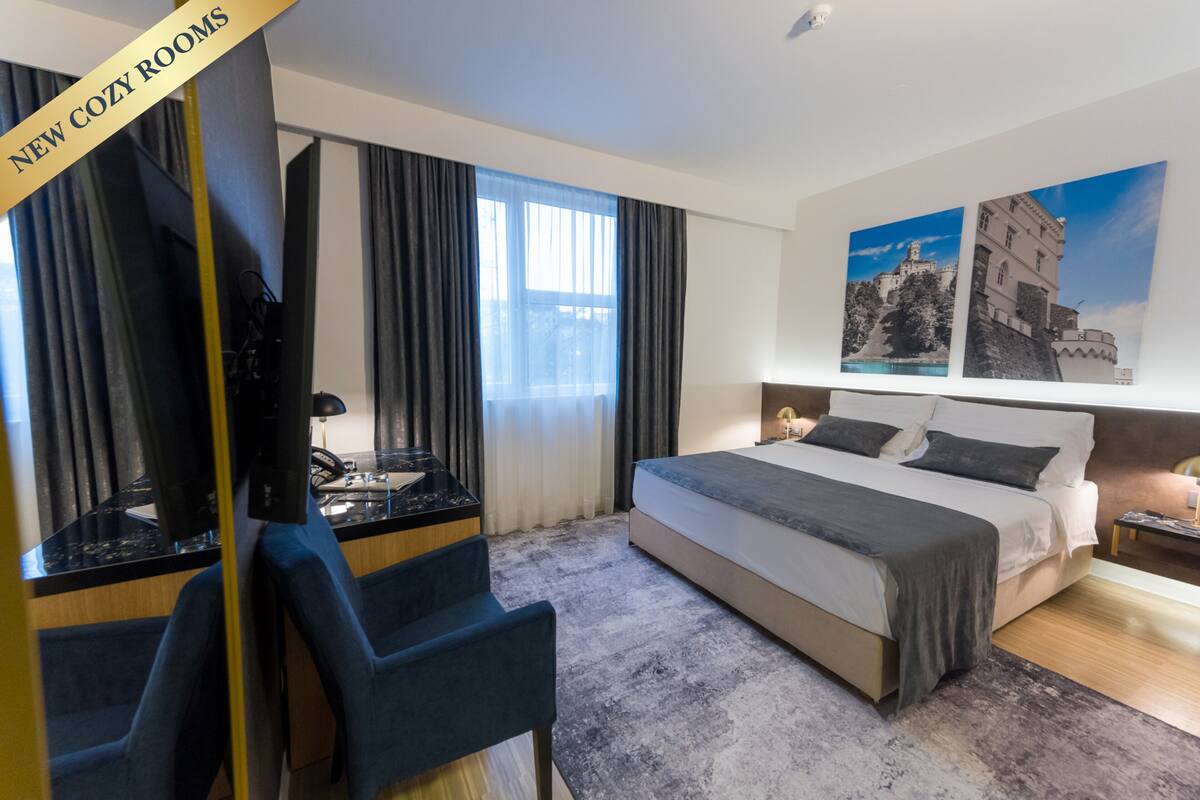 A hotel room with a bed, a desk with a chair, a TV, and large windows with curtains.