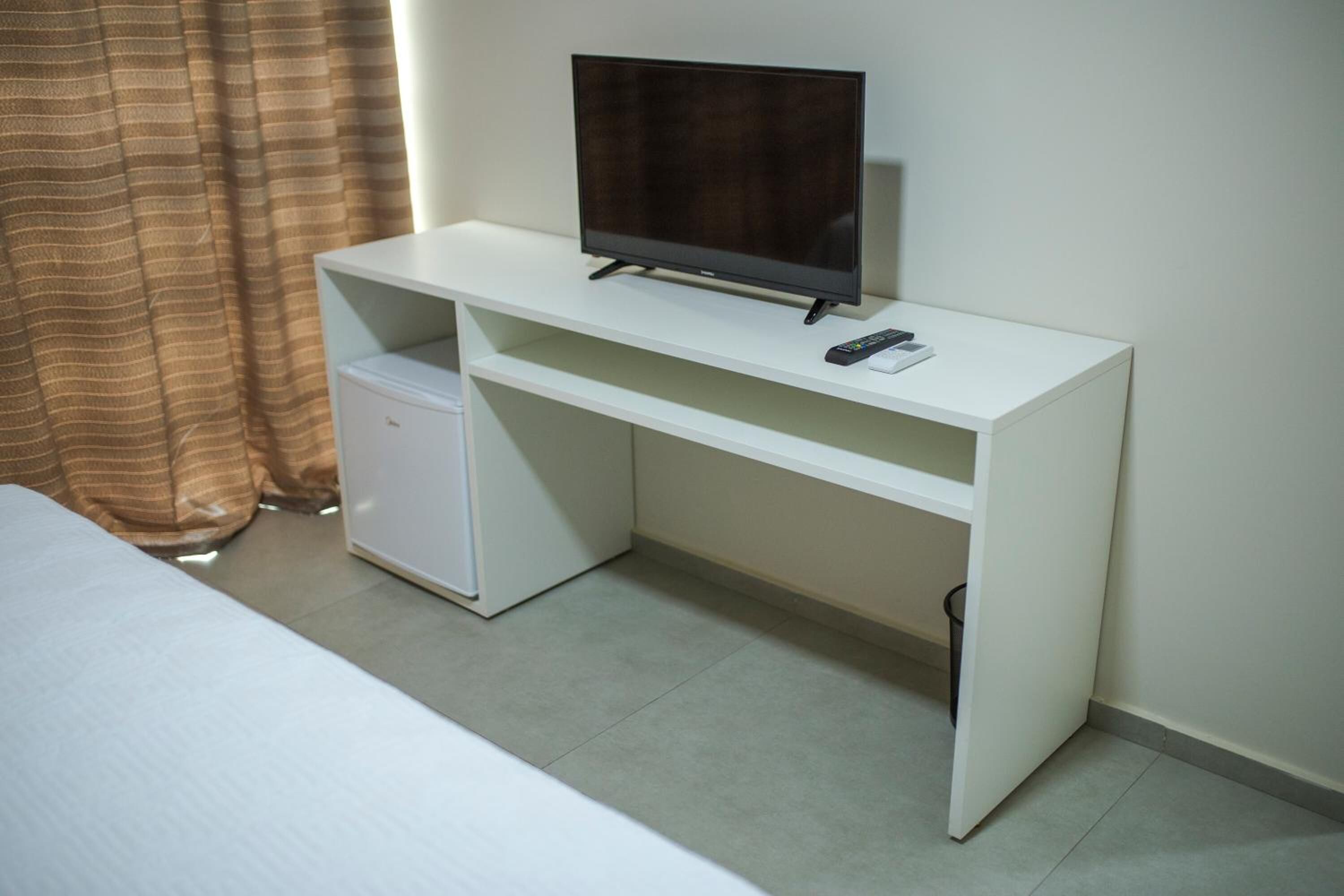 Una habitación de hotel con un escritorio blanco, televisor, refrigerador pequeño y una cama.