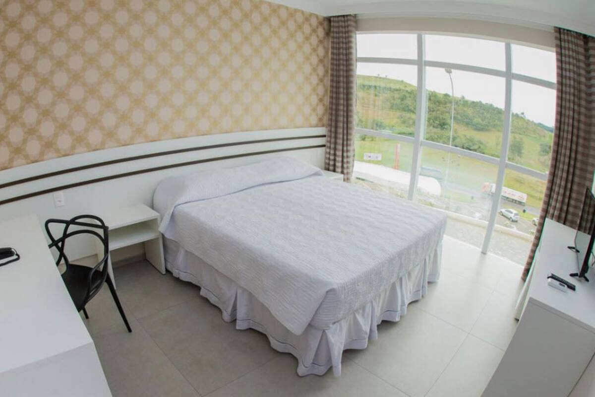 Un dormitorio con una cama grande, un escritorio y una silla. Hay una ventana con vistas a un paisaje verde.