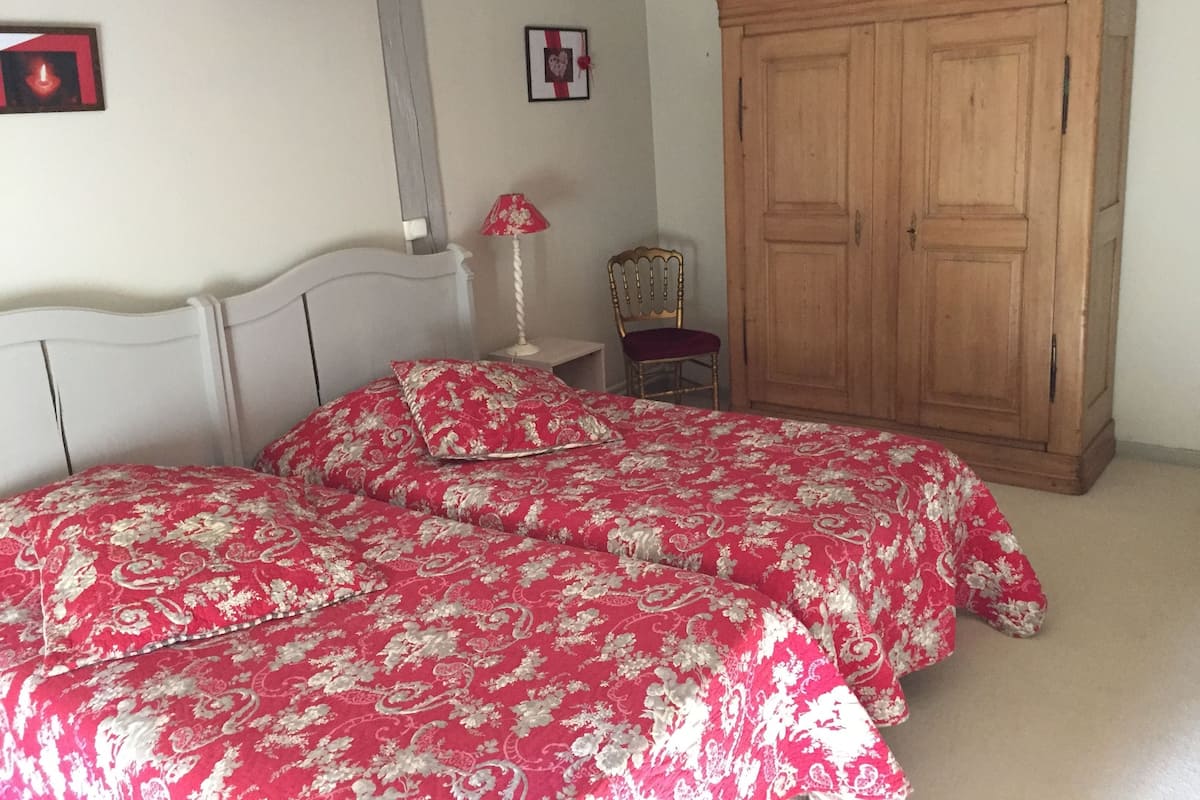 A bedroom with two beds, a wooden wardrobe, a chair, and a bedside table with a lamp.