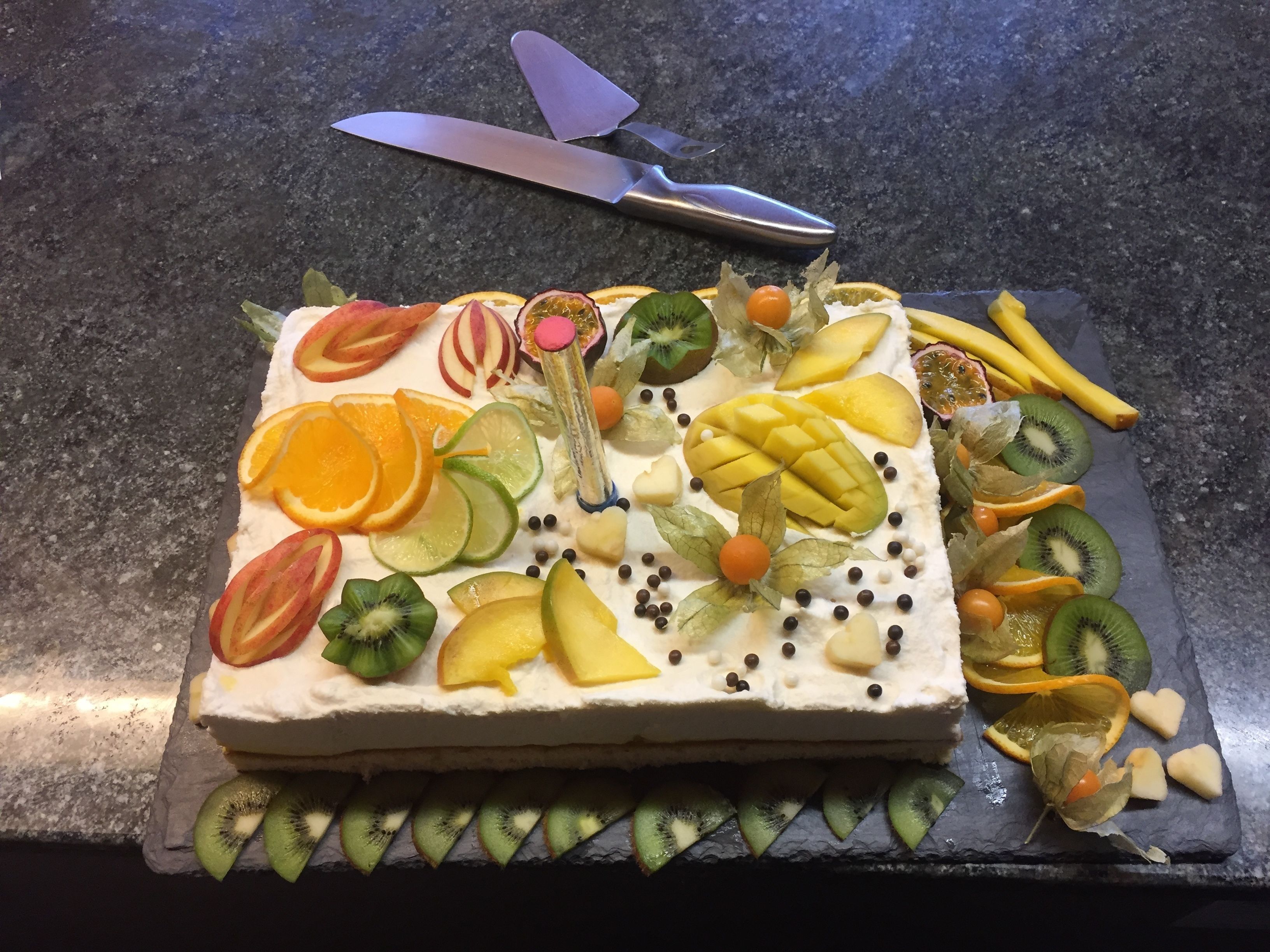 A square cake with various fruits and a knife on a dark countertop.