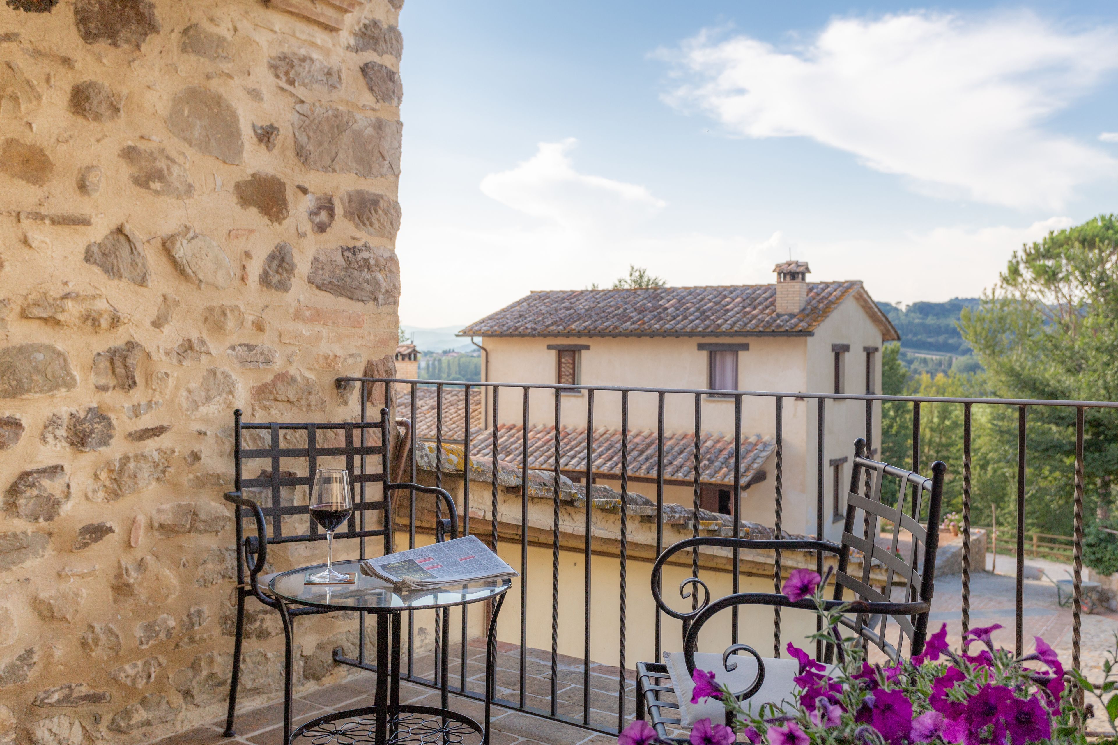 Un muro in pietra, un balcone con ringhiere in metallo, un tavolo con un bicchiere di vino e una sedia.