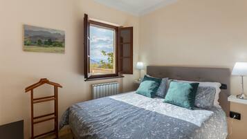 Quarto com cama, janela, radiador, mesa de cabeceira com abajur e um cabideiro de madeira.