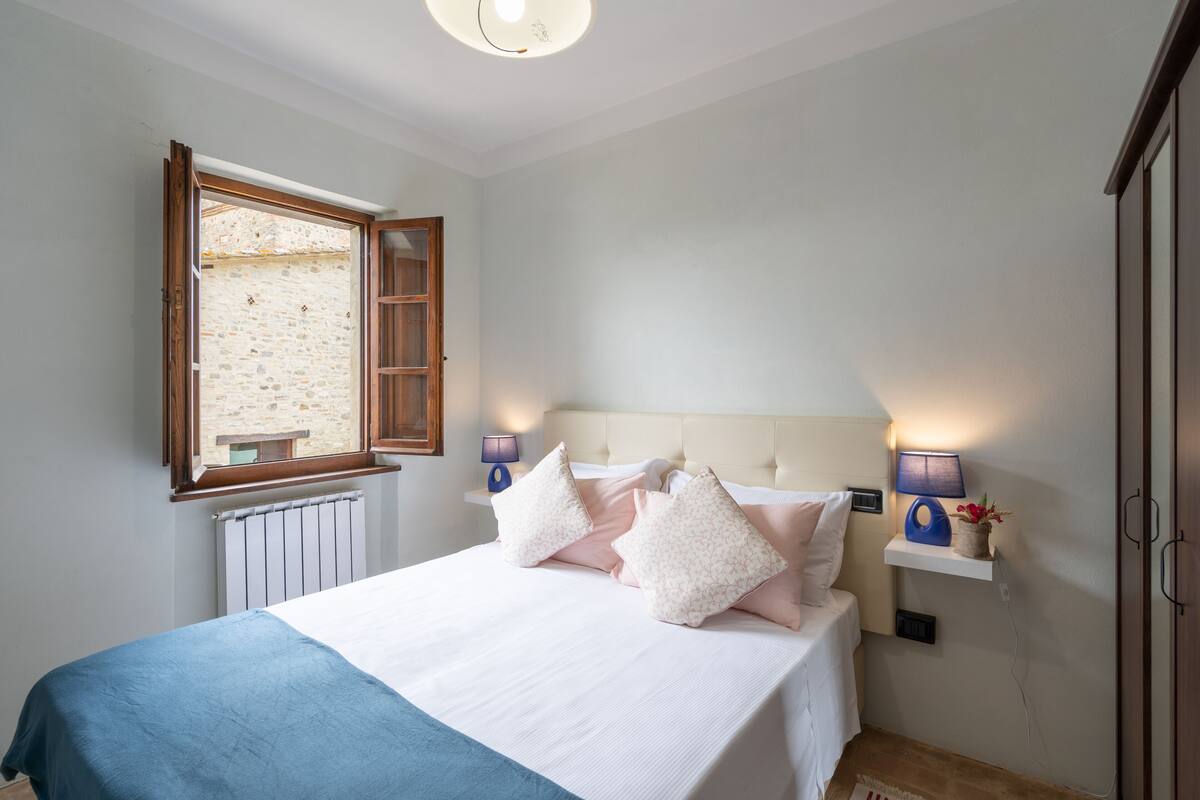 Een slaapkamer met een bed, kussens, een nachtkastje, een lamp, een raam en een radiator.