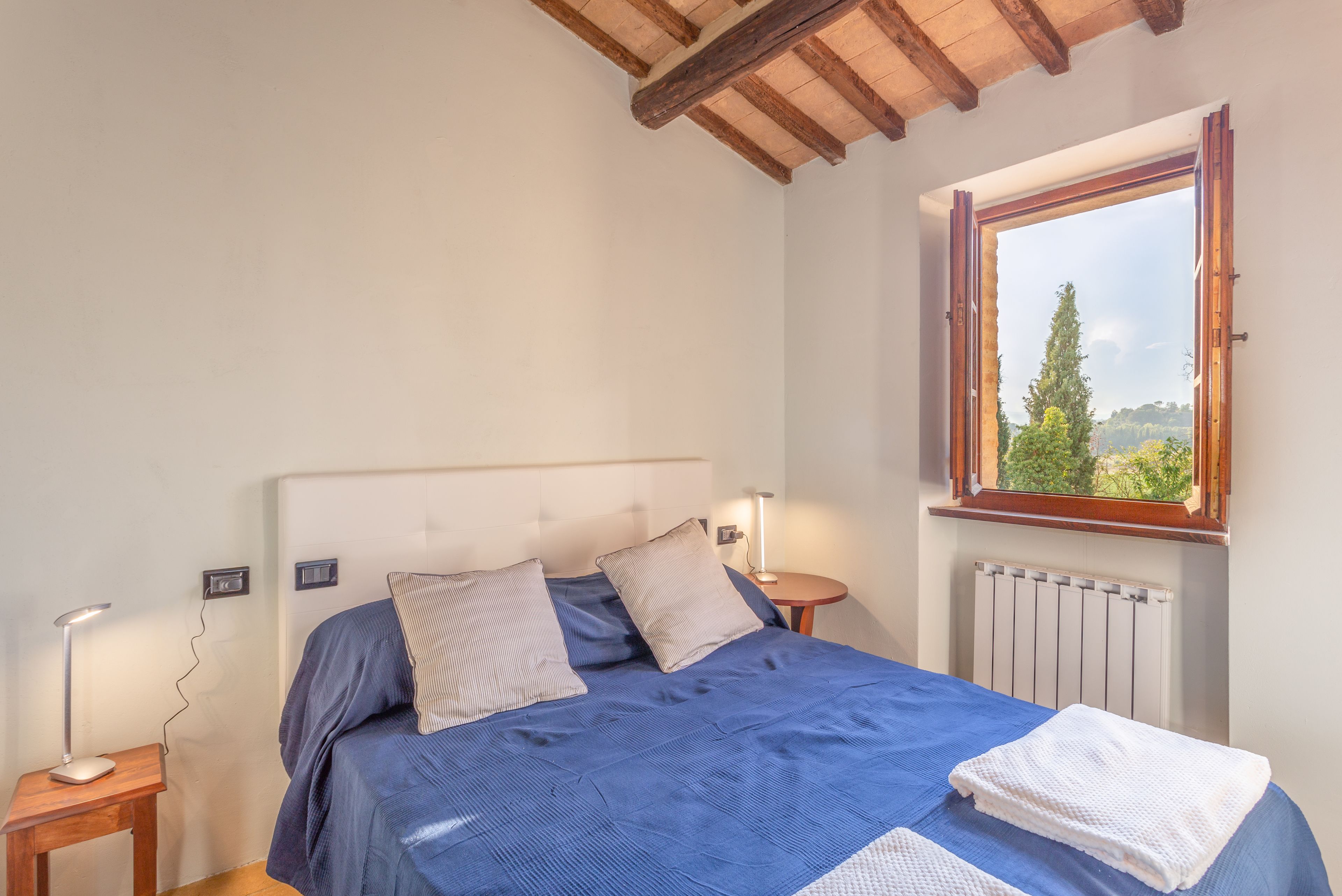 A bedroom with a bed, bedside lamps, a window with a view of trees, and a wooden ceiling.