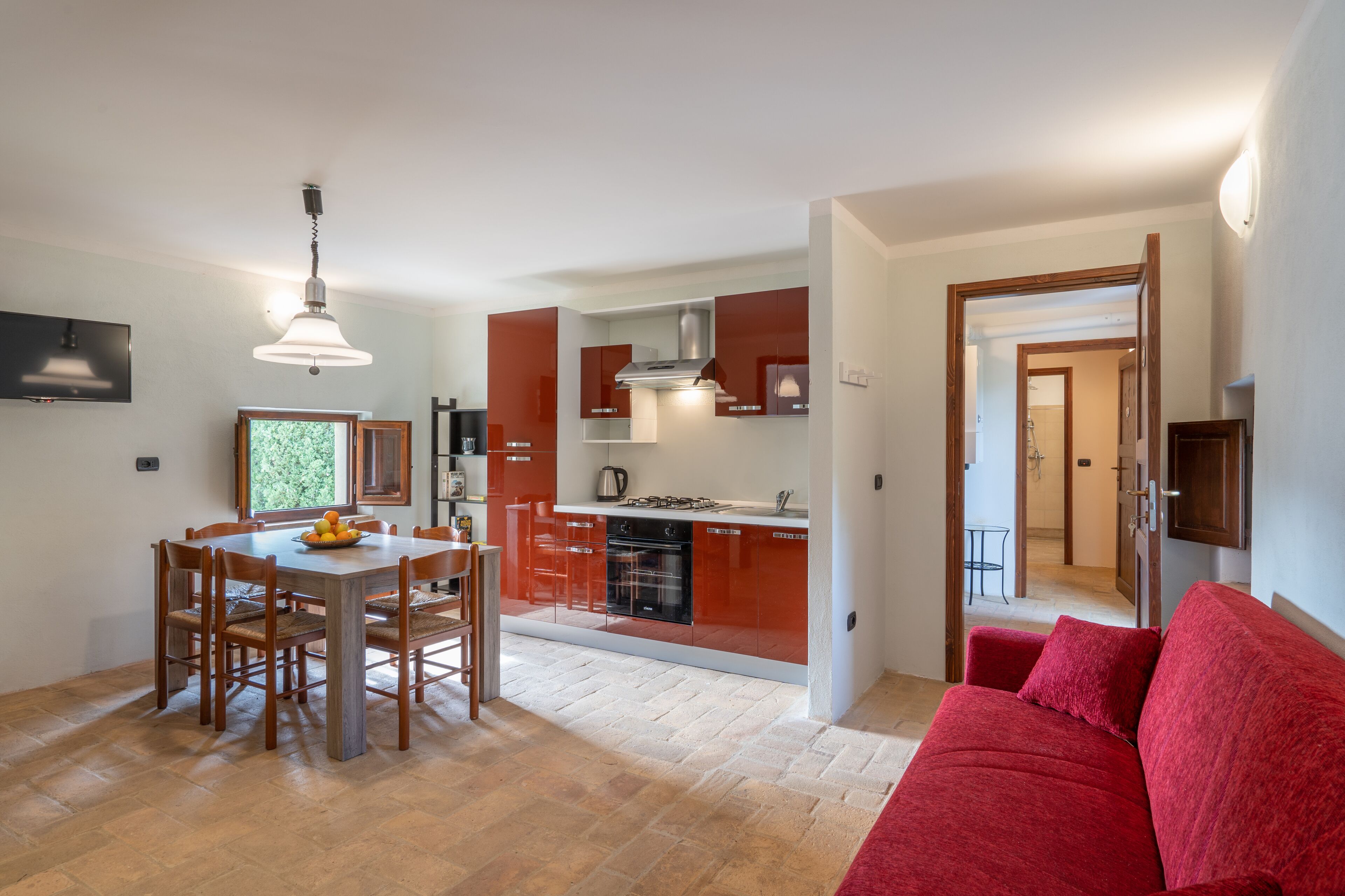 A modern kitchen with red cabinets, a dining area with wooden chairs, and a red sofa in a living room.
