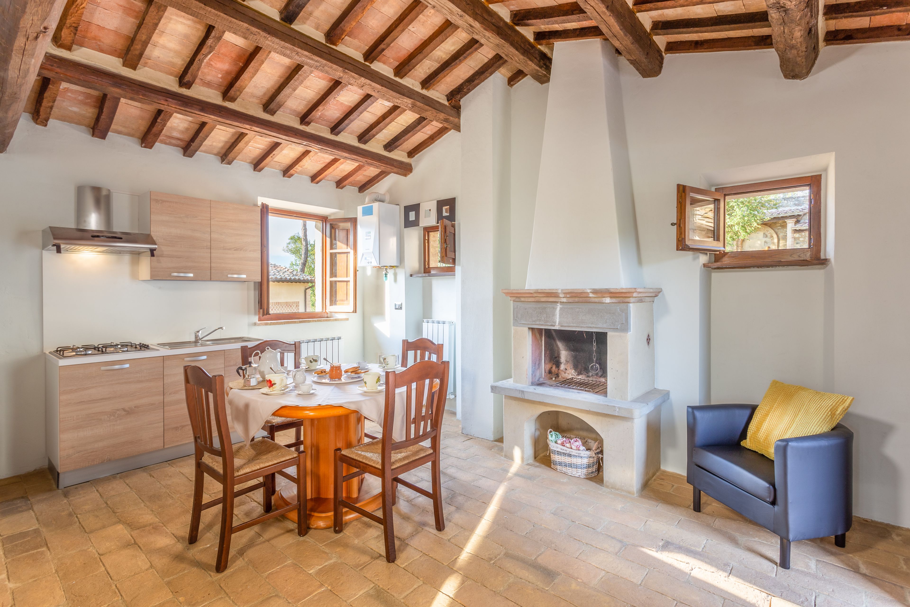 A cozy kitchen with wooden beams, a dining table with chairs, a fireplace, and a blue sofa with a yellow pillow.
