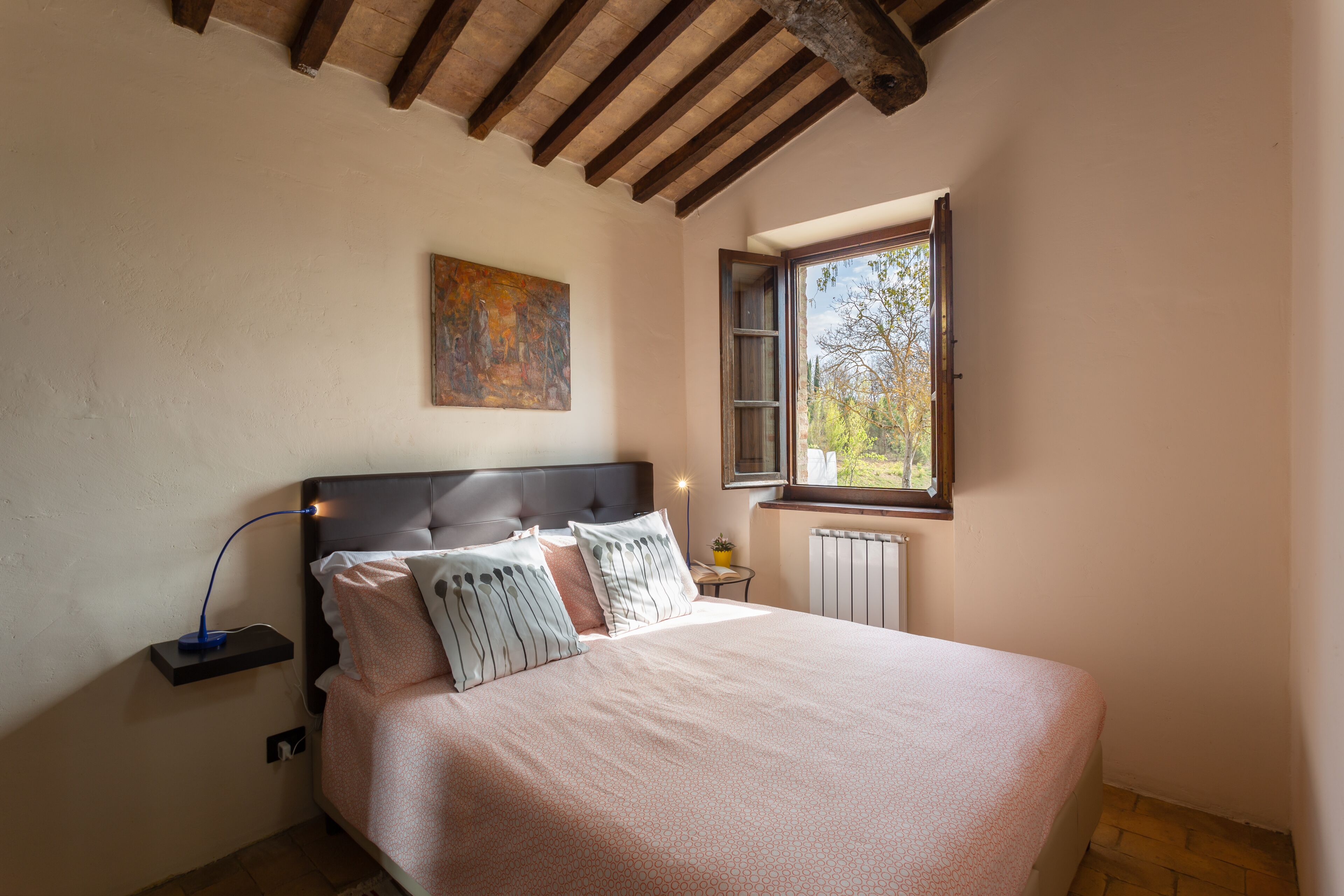 Un dormitorio con cama, mesita de noche, lámpara, ventana y un cuadro en la pared.