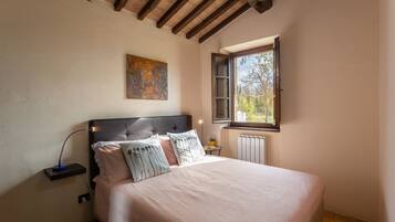 Un dormitorio con cama, mesita de noche, lámpara, ventana y un cuadro en la pared.