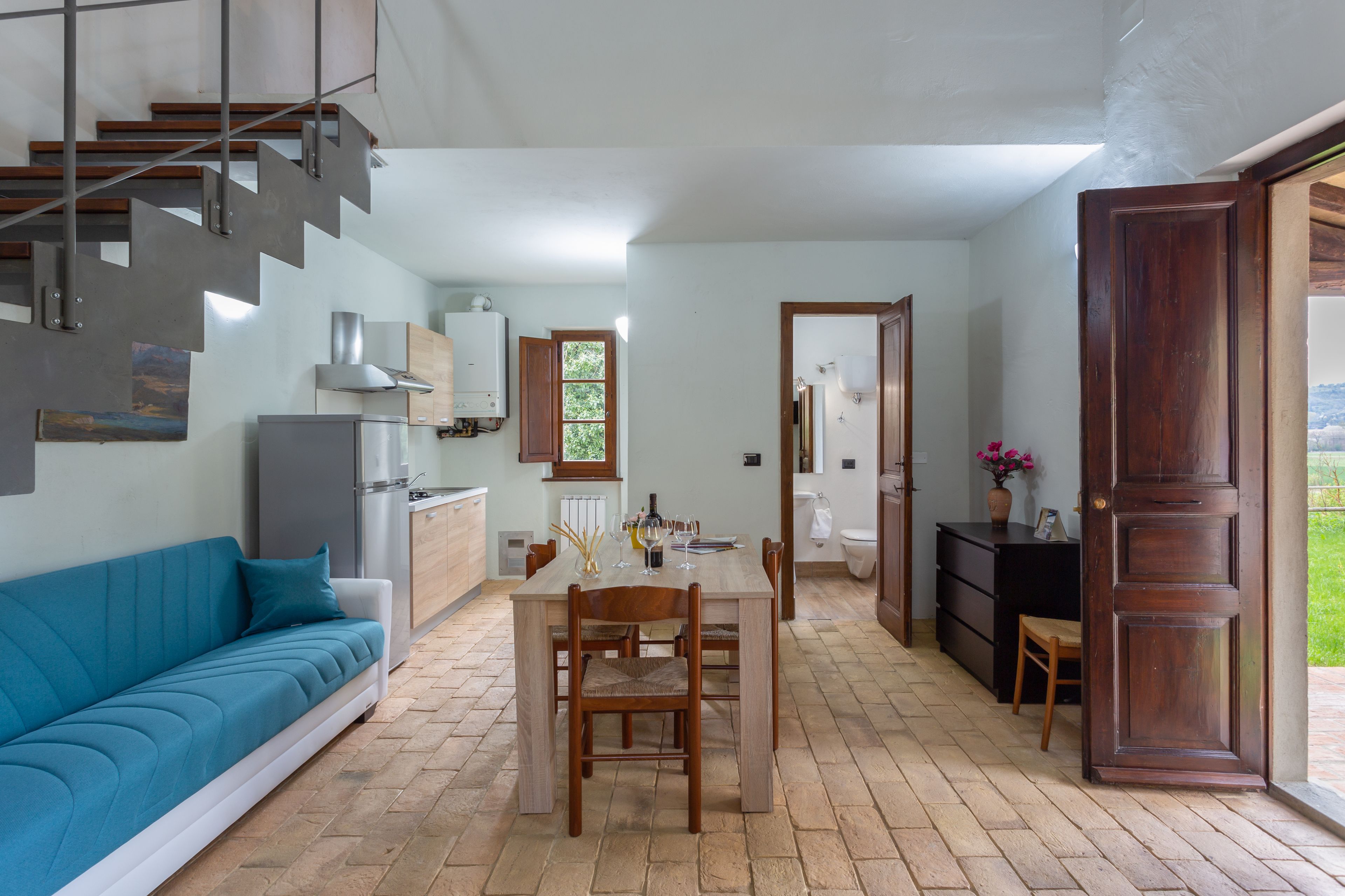 Una cocina moderna con horno empotrado, un comedor con mesa y sillas de madera, un sofá azul y una escalera que sube a otro nivel.